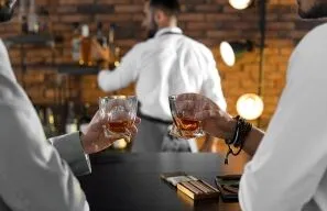 Two people toast with short whiskey glasses at a bar, bartender and warm brick wall in the background.