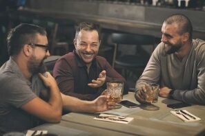Three men sit around a table at a bar, smiling and chatting while holding drinks.