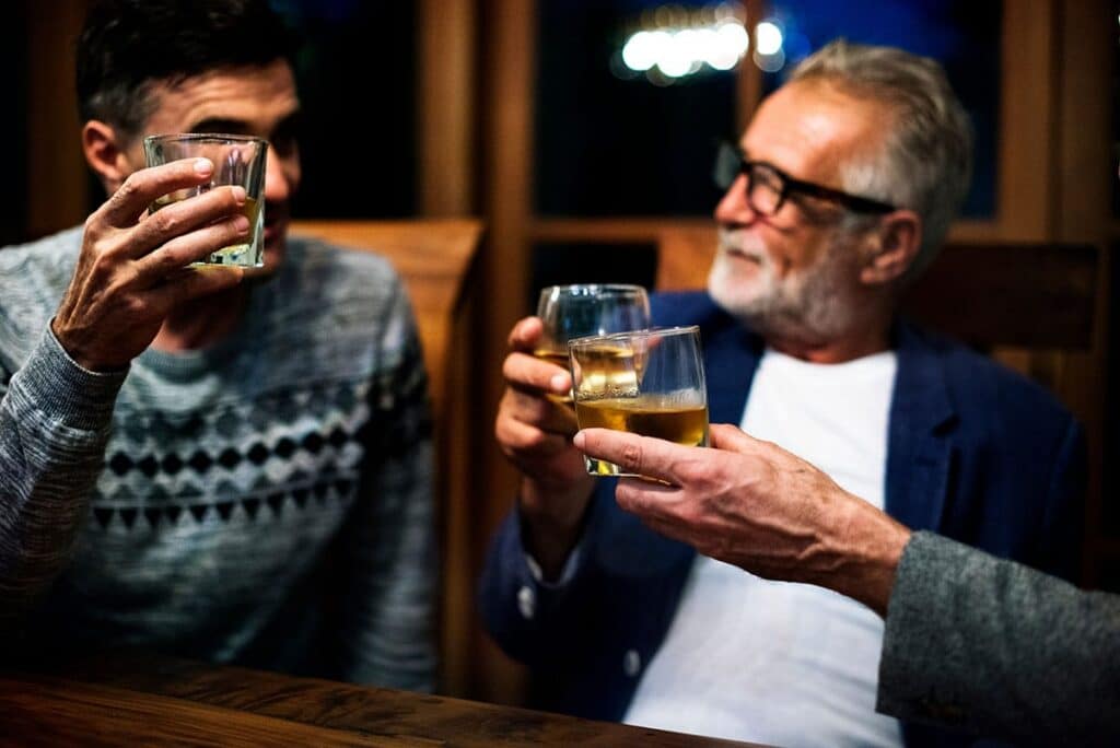 Three friends toast with whiskey glasses at a wooden bar in a warm, dimly lit setting.