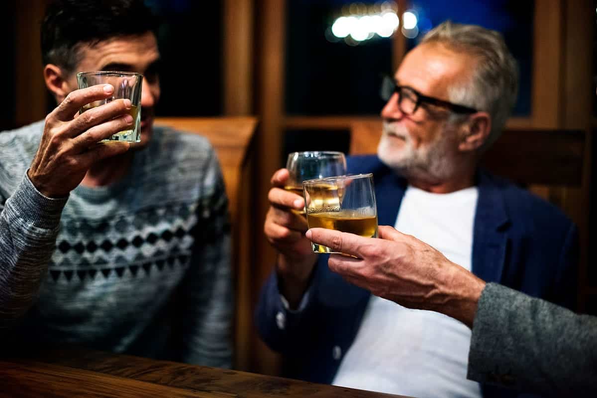 Three friends toast with whiskey glasses at a wooden bar in a warm, dimly lit setting.