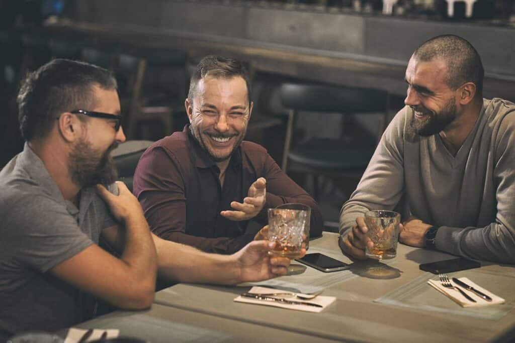 Three men sit at a bar table, laughing and talking over drinks in a casual social scene toasting with glasses and smiles.