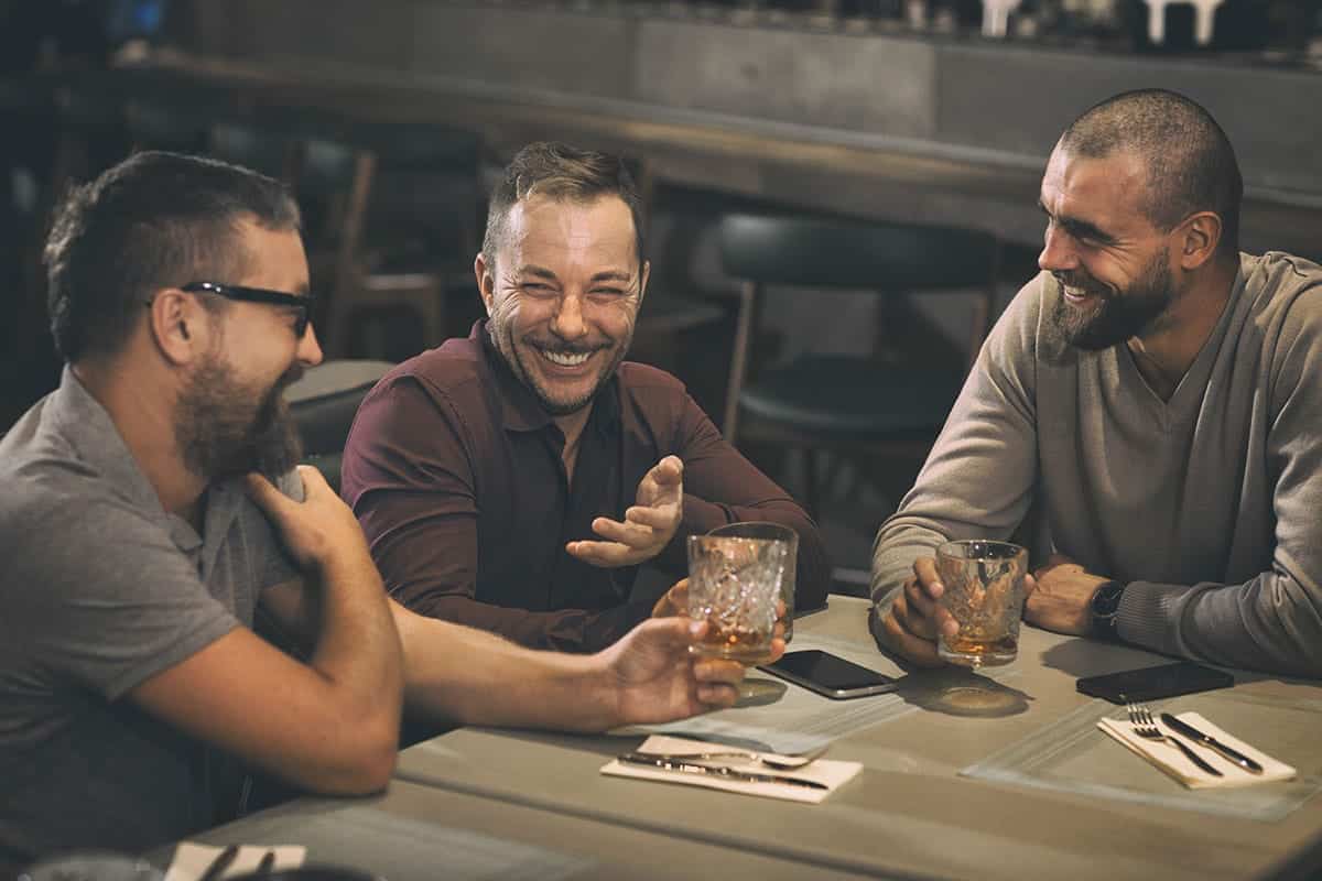 Three men sit at a bar table, laughing and talking over drinks in a casual social scene toasting with glasses and smiles.